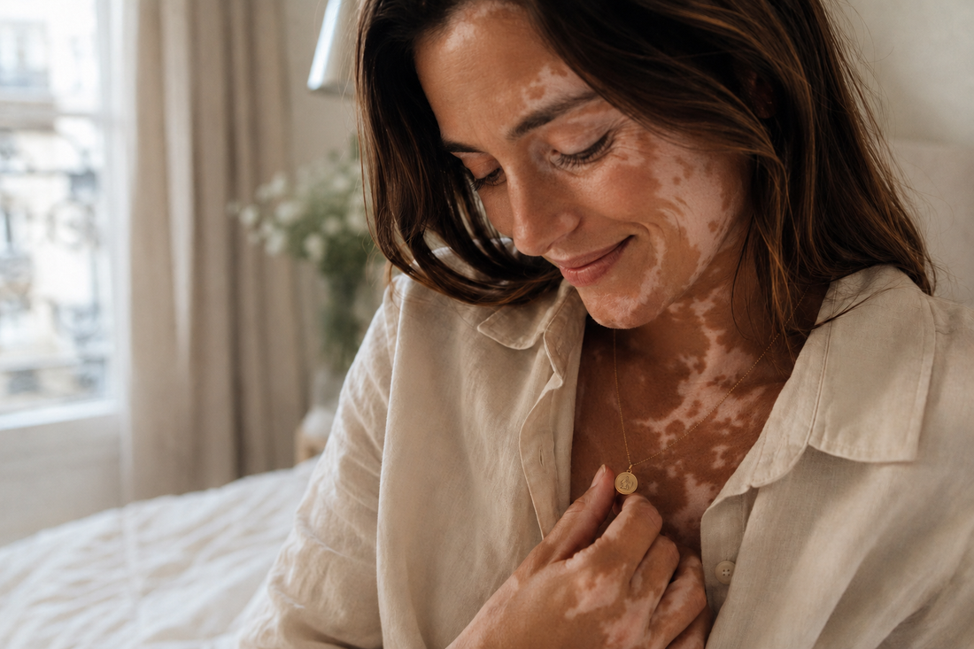Femme à la peau vitiligo portant un collier personnalisé doré, qu’elle touche doucement près du cœur dans une lumière naturelle, pour évoquer un bijou-souvenir intime et précieux.